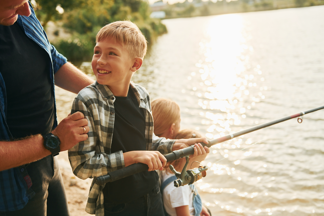 Smiling boy fishing at a pond with his family at sunset, enjoying a bonding moment and learning to fish in a well-stocked pond.